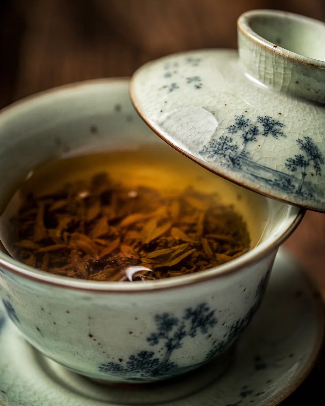 A blue-and-white gaiwan holding steeped leaf and pale gold liquor, its lid resting across the rim.