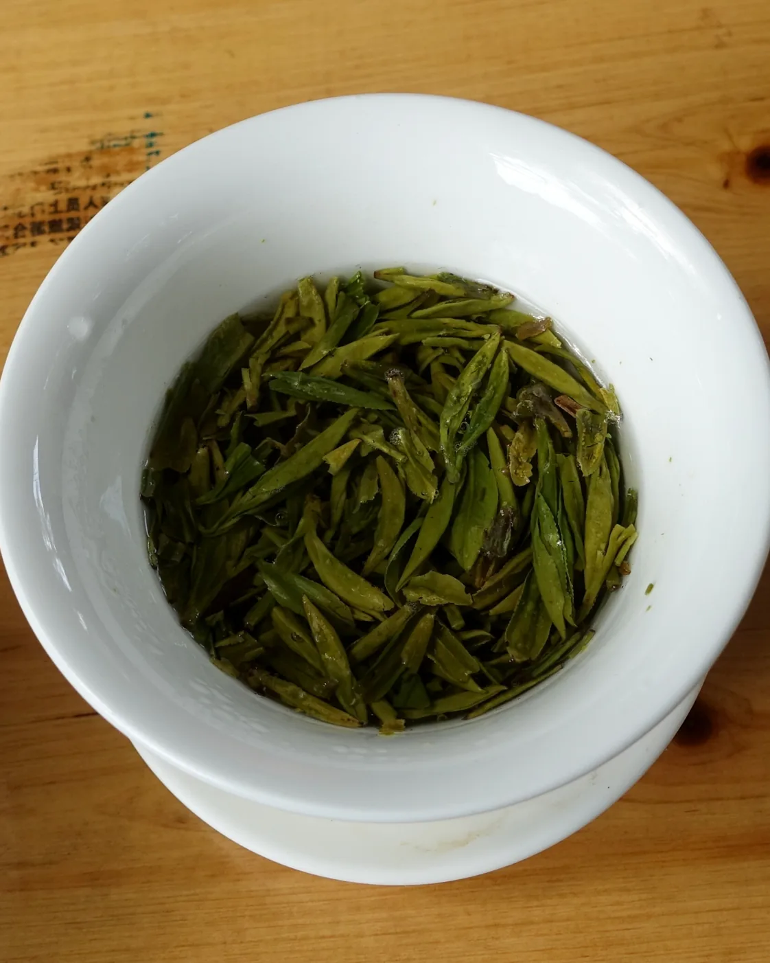 Whole green tea leaves steeping in a white gaiwan on a wooden teahouse table.