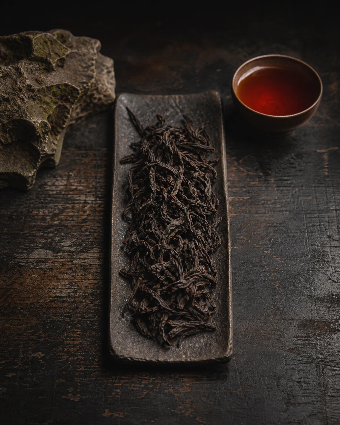 Da Hong Pao rock oolong: long twisted charcoal-dark leaves on a stone slab beside a cup of deep amber liquor.