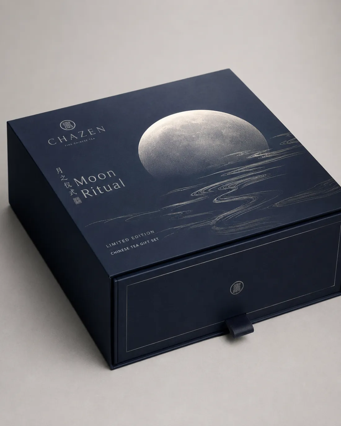 Closed navy drawer box printed with a large silver moon above stylised silver water, with a ribbon pull at the base