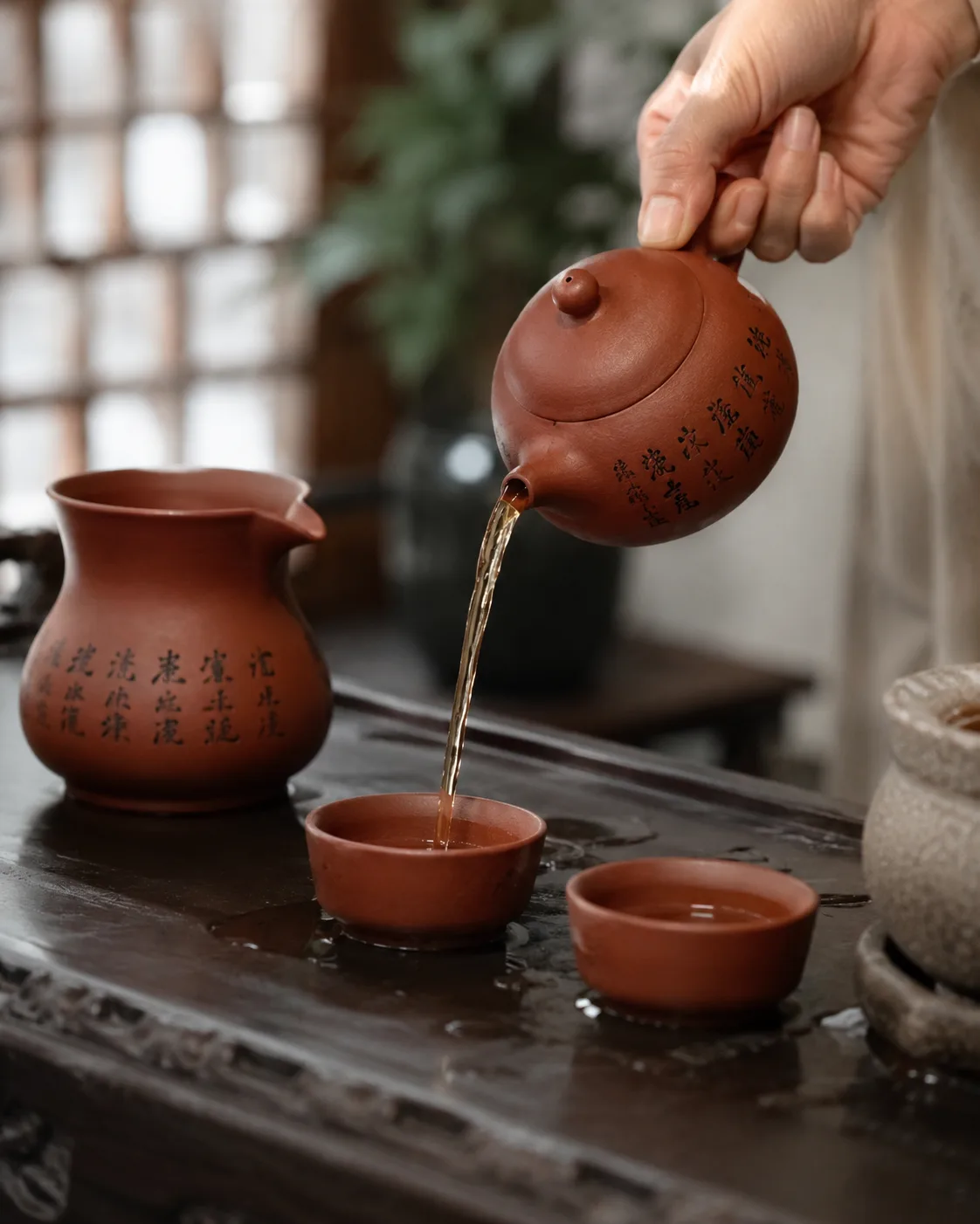 A hand tips a small clay teapot into two cups, beside a clay pitcher carved with characters.