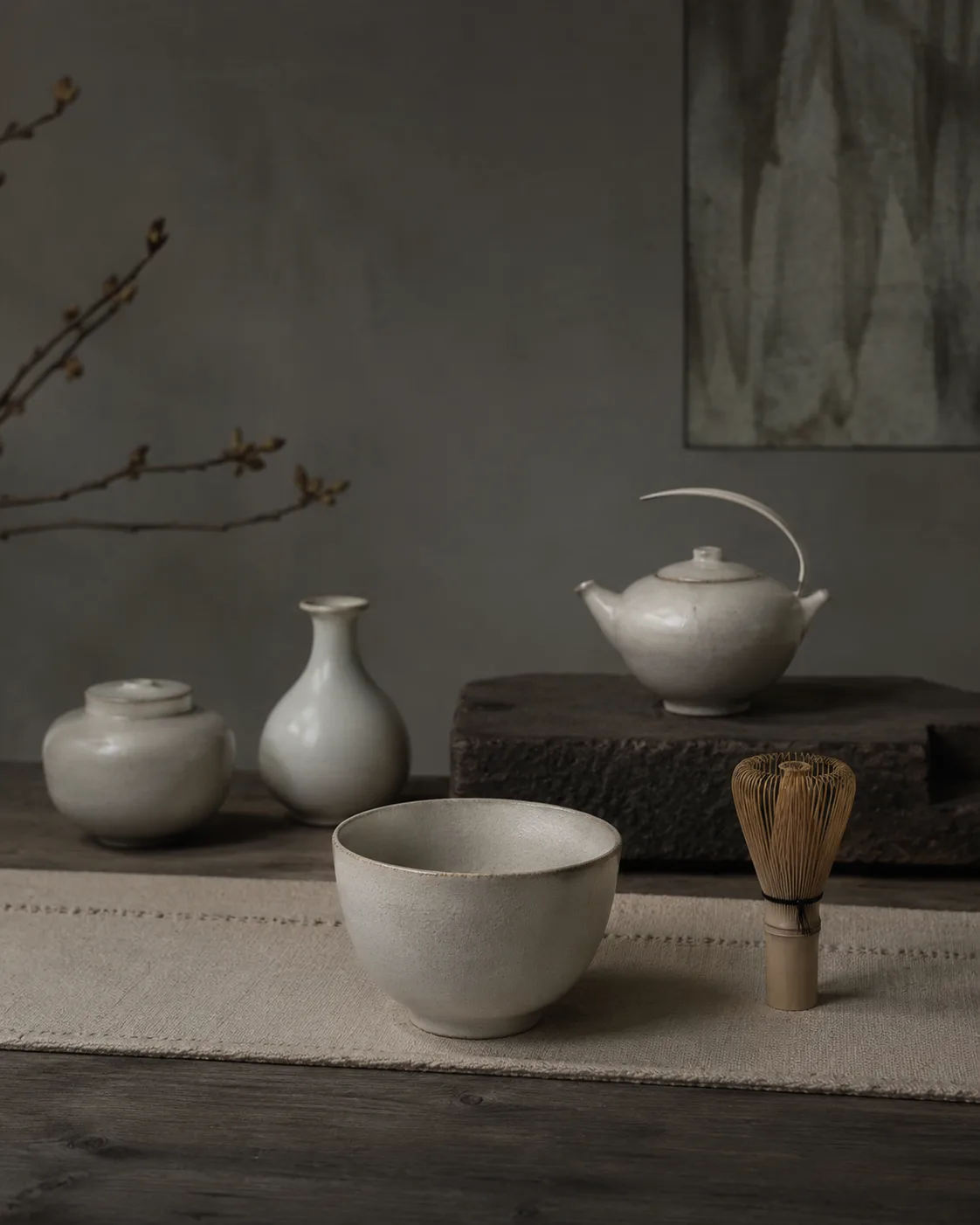 Pale white vessels — a lidded jar, a bottle vase, a bowl and a small pot — with a bamboo whisk on a linen cloth.