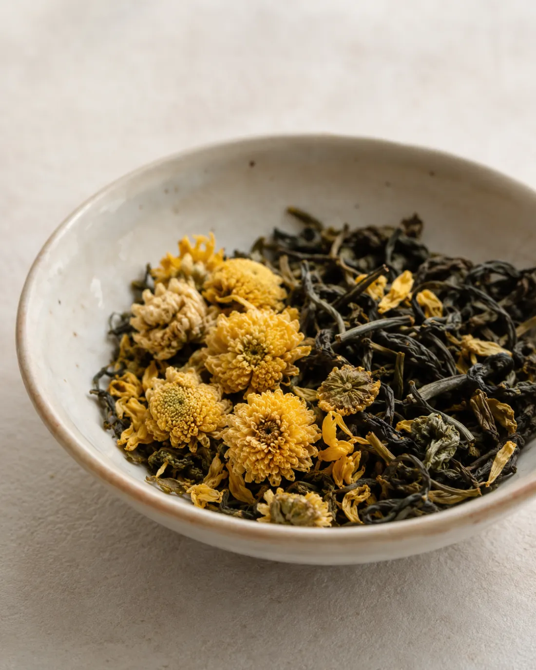Dried chrysanthemum heads mixed through dark green tea leaf in a shallow stoneware dish.